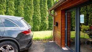 En bil som laddas vid en laddstation med en träbyggnad och en grön häck i bakgrunden.