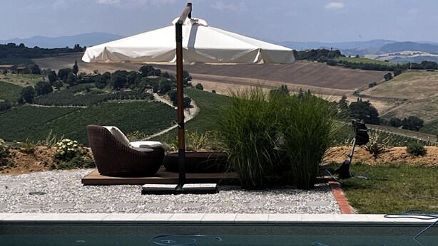 A pool with a chair and umbrella, surrounded by a gravel area and greenery, with a view of rolling hills in the background.