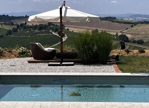 Una piscina con una silla y una sombrilla, rodeada de grava y follaje, con vistas a colinas onduladas al fondo.