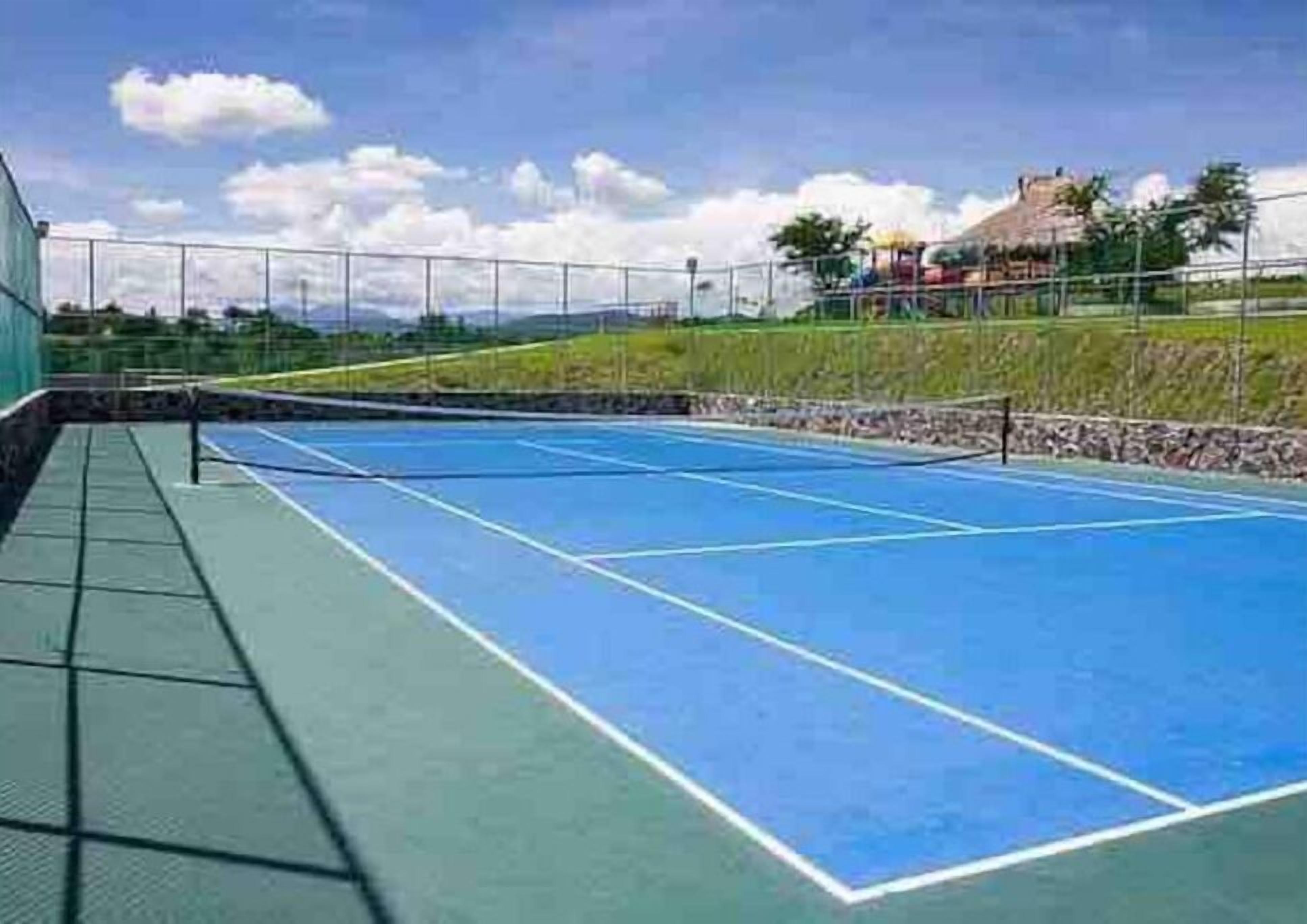 An outdoor tennis court with a green and blue surface, surrounded by a fence and a grassy area with a building in the background.