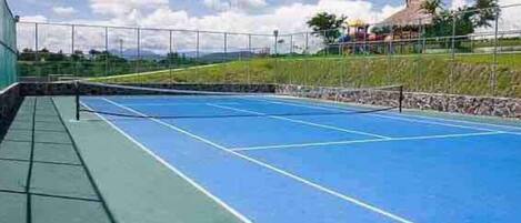 An outdoor tennis court with a green and blue surface, surrounded by a fence and a grassy area with a building in the background.