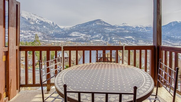 A round table with a patterned tabletop, surrounded by metal chairs, on a balcony with a view of snow-capped mountains and a body of water. - Les Fauvettes - appartement vue imprenable sur montagne (Embrun)