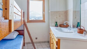 A small, compact room with a bunk bed, a window with a mountain view, a sink with a mirror, and wooden cabinets. - Les Fauvettes - appartement vue imprenable sur montagne (Embrun)