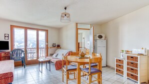 A spacious room with a dining table, chairs, a sofa, a TV, and a large window with a view of snow-capped mountains. - Les Fauvettes - appartement vue imprenable sur montagne (Embrun)