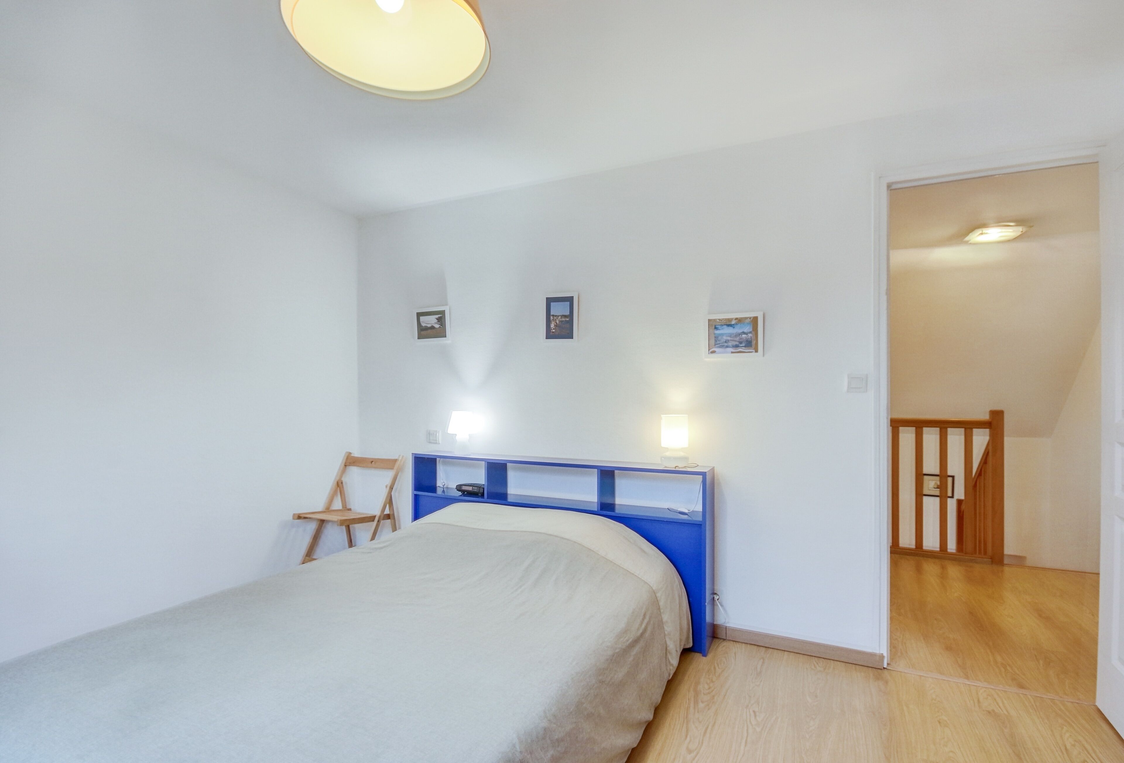 Een slaapkamer met een bed, een blauw hoofdeinde, een houten stoel en een trap naar een andere kamer.