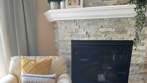 A living room with a stone fireplace, a white armchair with a 'Farmhouse' pillow, a small green plant, and a vase with yellow flowers.