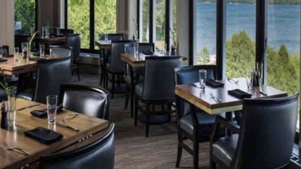 A restaurant with large windows offering a view of a lake. Tables are arranged with chairs, and there are glasses and napkins on the tables.