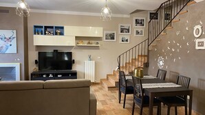 Ett vardagsrum med en soffa, en tv och en matplats med stolar och ett bord. Det finns en trappa med ett räcke och en vägg som är dekorerad med bilder och fjärilar.