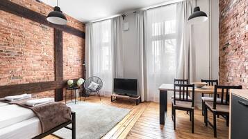Habitación moderna con pared de ladrillo, suelo de madera, cama, televisor, mesa de comedor con sillas y una mesita auxiliar con un florero.