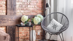 Una habitación con pared de ladrillo, una silla negra con almohada de estrella, una mesita con un florero y una lámpara.