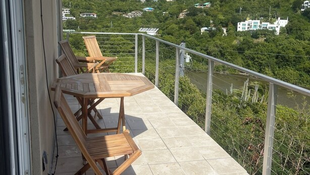 A balcony with wooden chairs and a table, overlooking a hillside with scattered buildings. - Breezy Hilltop, Hart Bay View, only minutes from town (Cruz Bay)