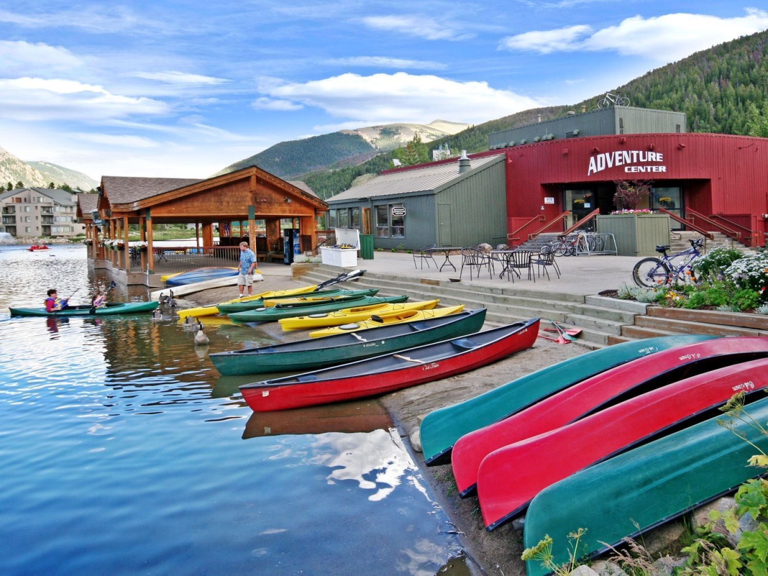 Una cabaña junto al lago con canoas amarradas en la orilla, un edificio de madera con un porche, y un edificio rojo que dice "Adventure Center".