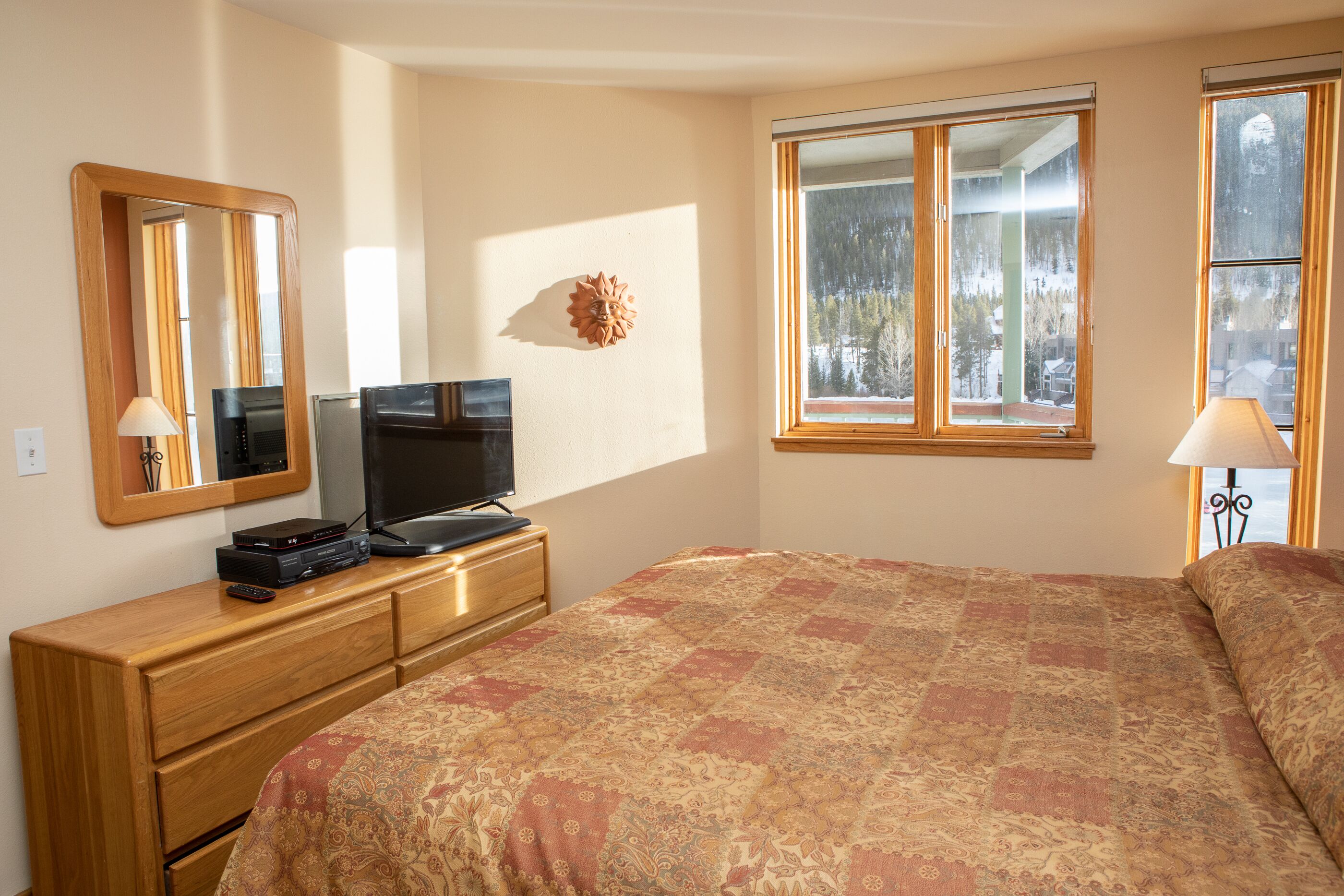 A bedroom with a bed, a dresser with a television, a mirror, and a window with a view of trees.