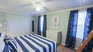Quarto com cama, penteadeira, ventoinha no teto e cortinas com padrão de âncoras.