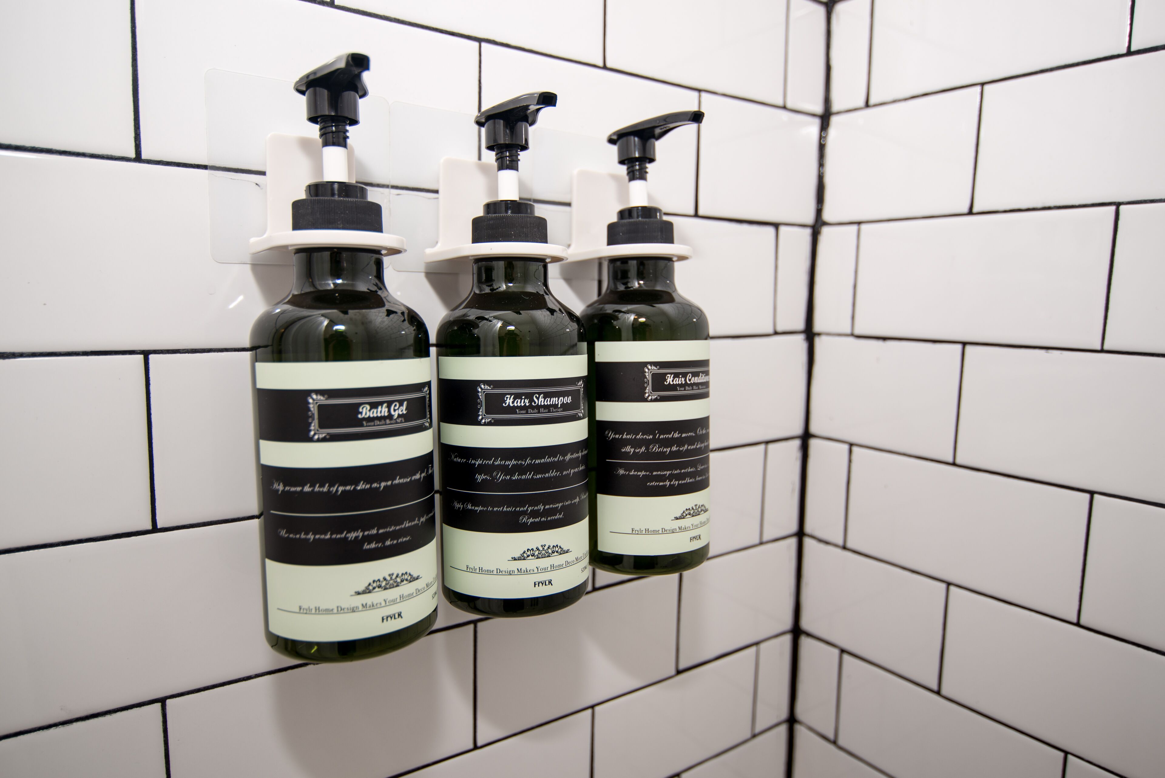 Three bottles of bath oil and hair shampoo on a tiled wall.