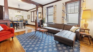 A living room with a brown sofa, red armchair, and a blue patterned rug. There is a dining area in the background with a table and chairs.