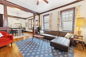 A living room with a brown sofa, red armchair, and a blue patterned rug. There is a dining area in the background with a table and chairs.