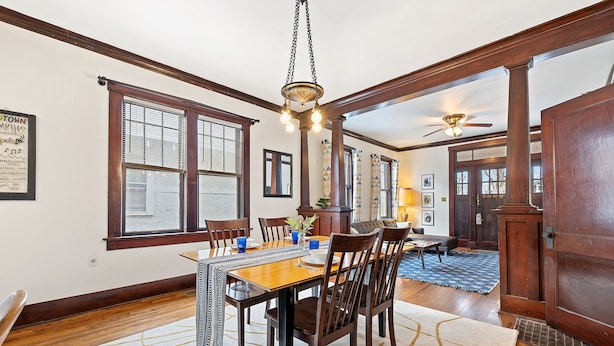 A dining room with a wooden table, chairs, and a chandelier. There are windows with blinds, a ceiling fan, and a rug on the floor.