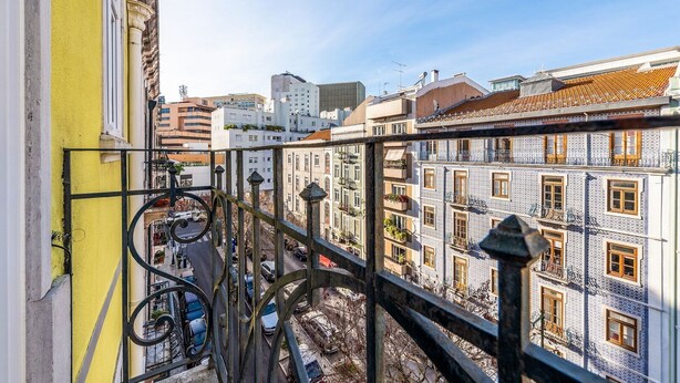 Un balcon doté d’une balustrade en métal, offrant une vue sur un paysage urbain avec des bâtiments à l’architecture traditionnelle et un ciel dégagé.