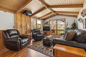 Un salón acogedor con vigas de madera, sofás de cuero, un televisor y un ventanal con vistas al exterior.