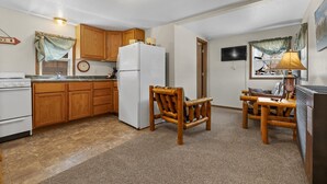 Una cocina compacta con armarios de madera, un refrigerador blanco, una mesa de comedor con sillas, un televisor y una lámpara.