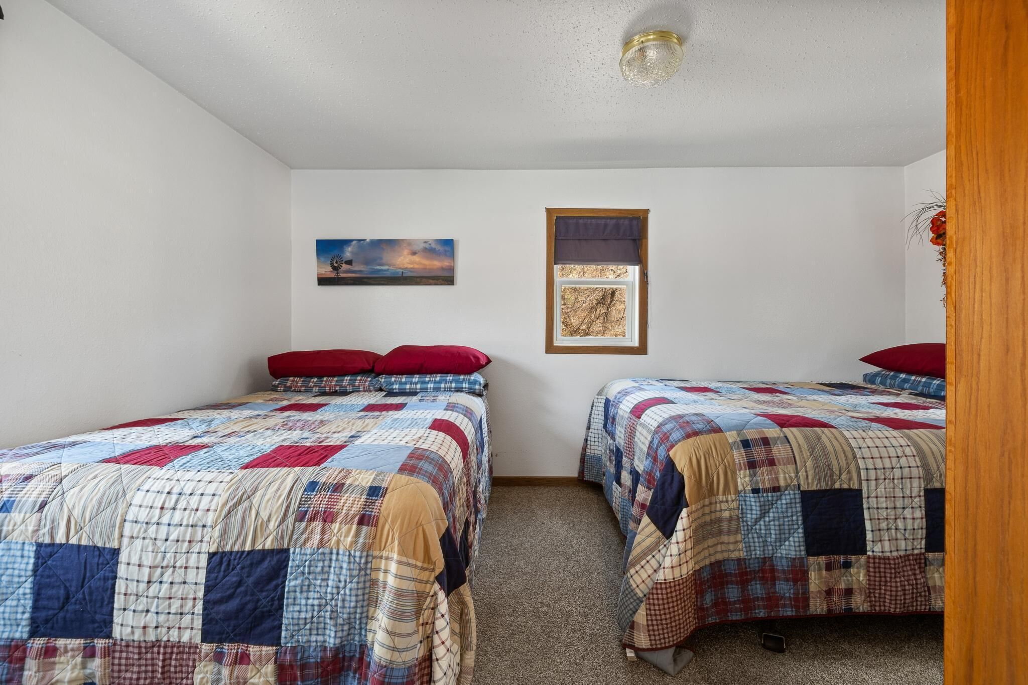 A bedroom with two beds, each with a quilted bedspread. There is a framed picture on the wall and a window with a curtain.