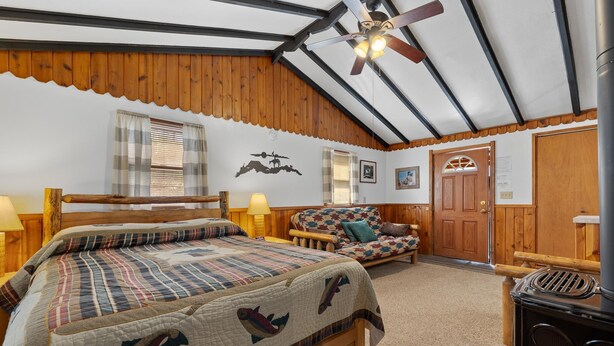 Une chambre confortable avec un lit, une tête de lit en bois, un canapé, un ventilateur de plafond et un poêle à bois.
