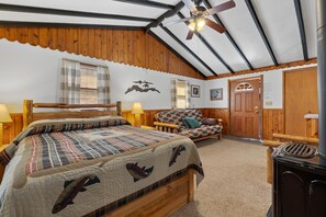Quarto aconchegante com cama, cabeceira de madeira, sofá, ventilador de teto e lareira a lenha.