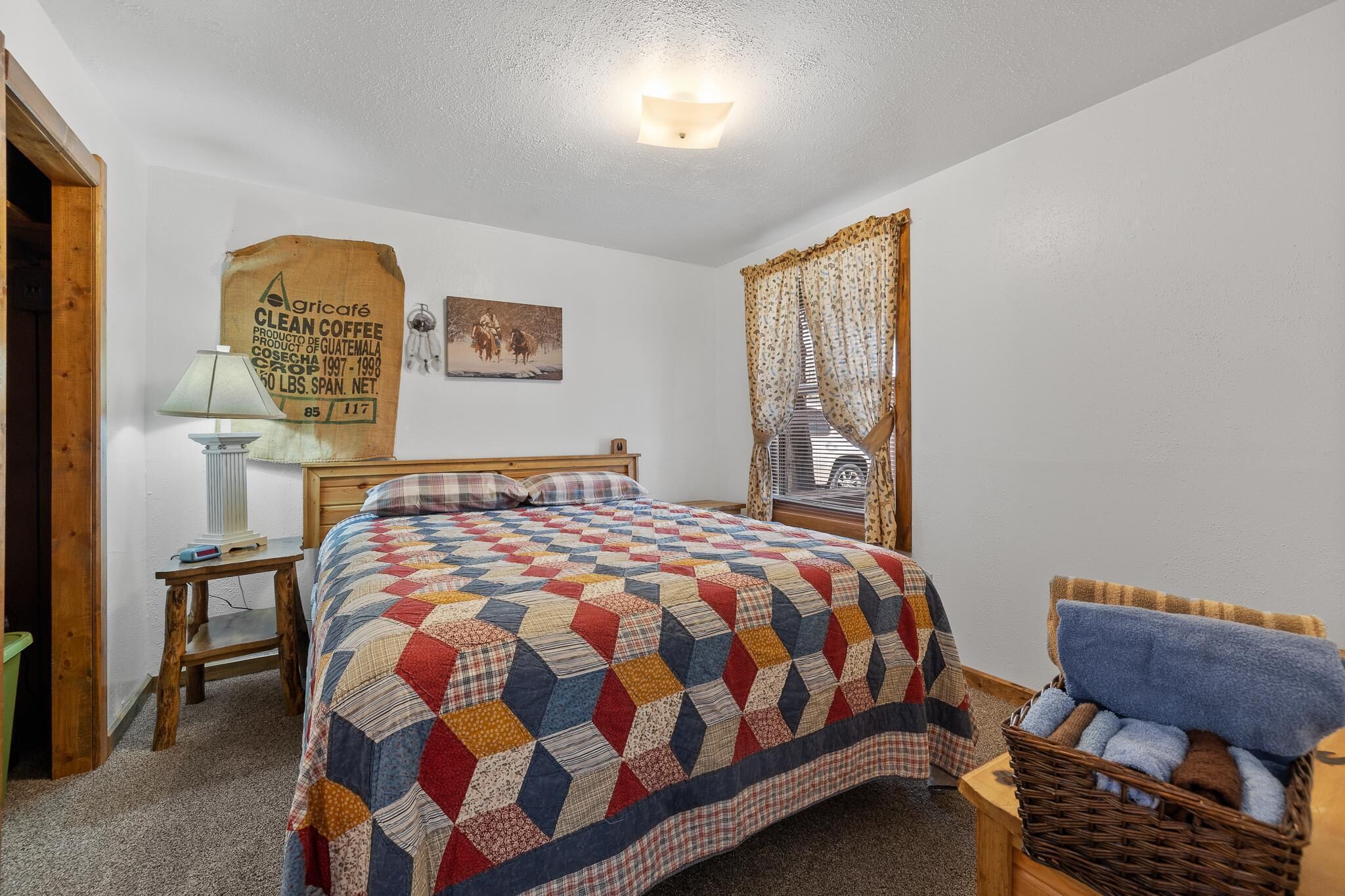 Une chambre avec un lit, une lampe, un panier contenant des serviettes et un mur orné d’un sac de café.