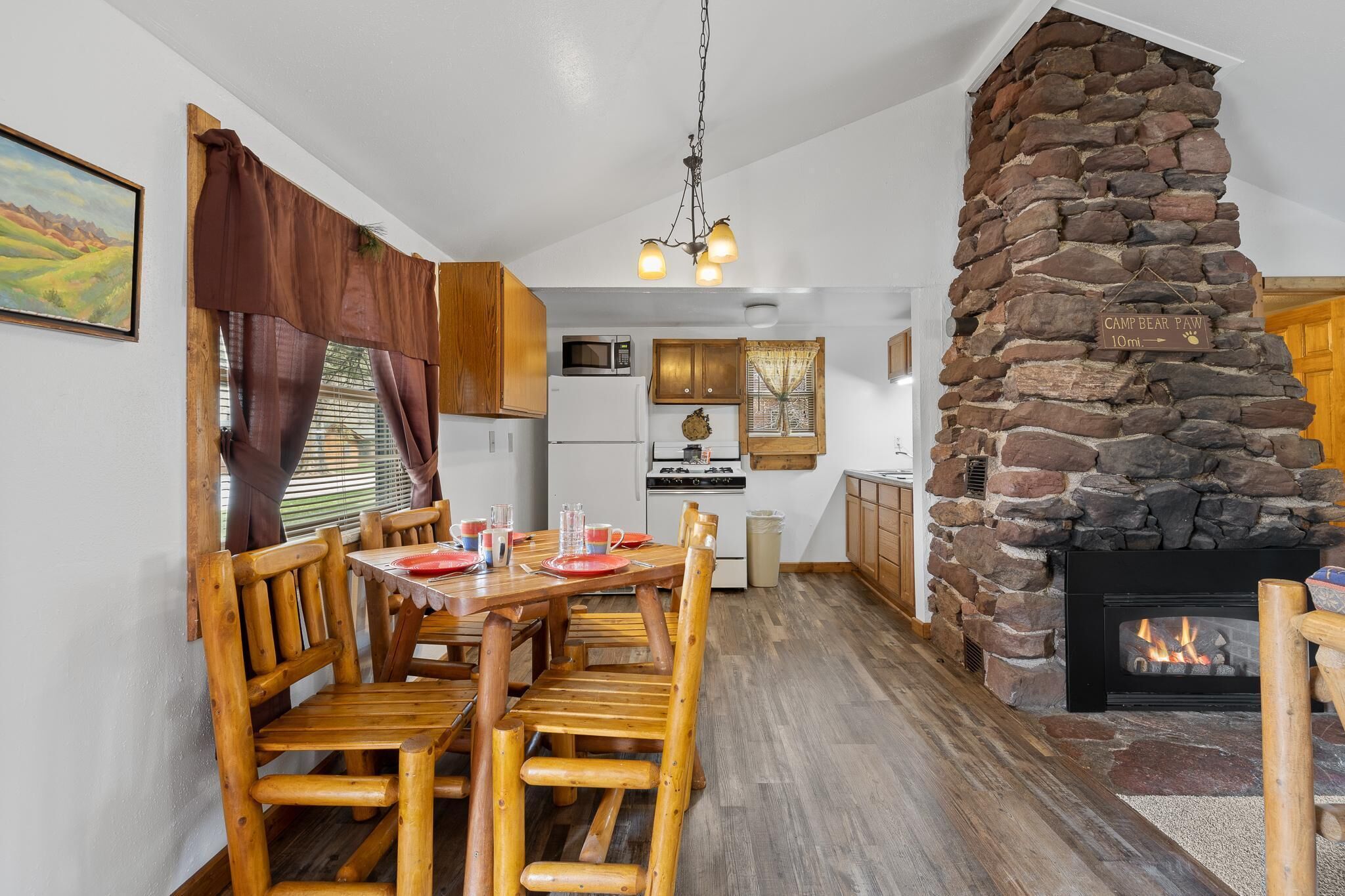 Un coin repas chaleureux avec une table et des chaises en bois, une cheminée en pierre et une cuisine en arrière-plan.