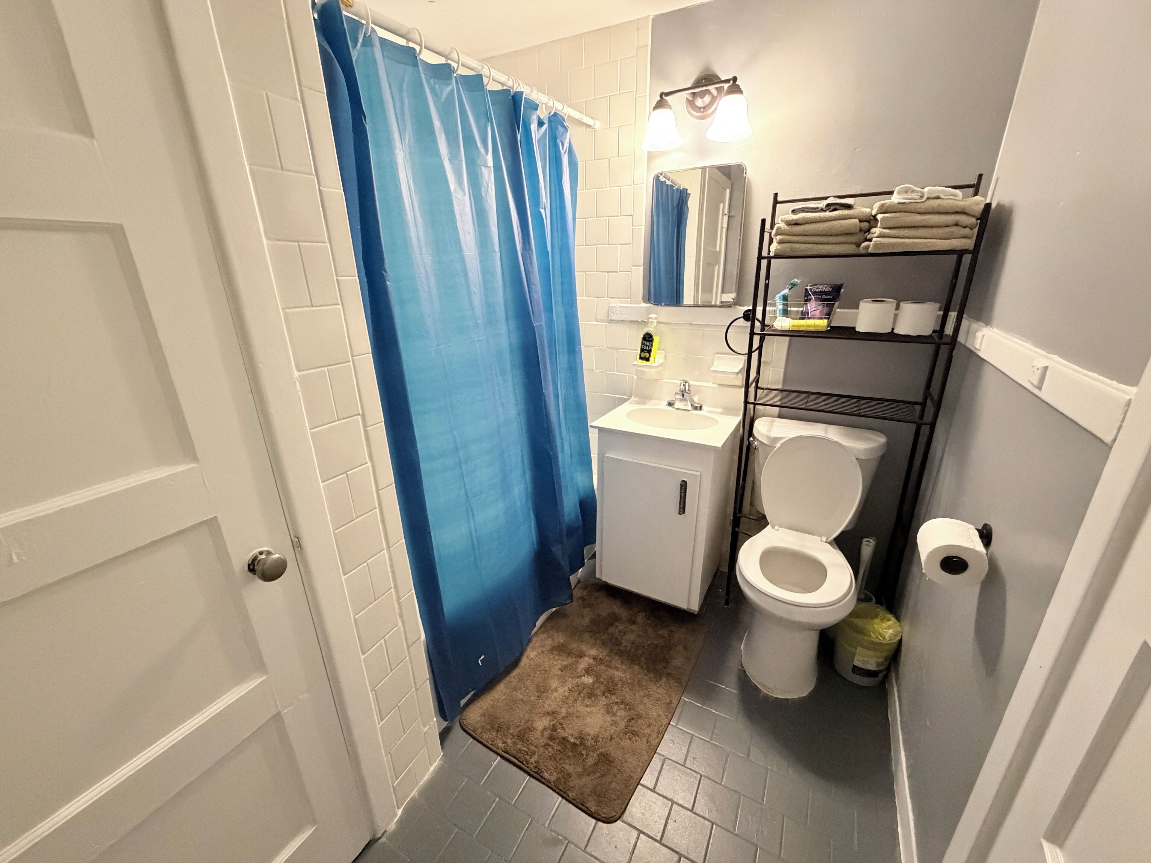 A small bathroom with a white sink, toilet, and blue shower curtain. There is a mirror above the sink and a shelf with towels and toiletries.