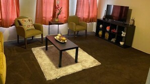 Uma sala de estar com duas poltronas, uma mesa de centro, um rack para TV com objetos decorativos e uma janela com cortinas vermelhas.