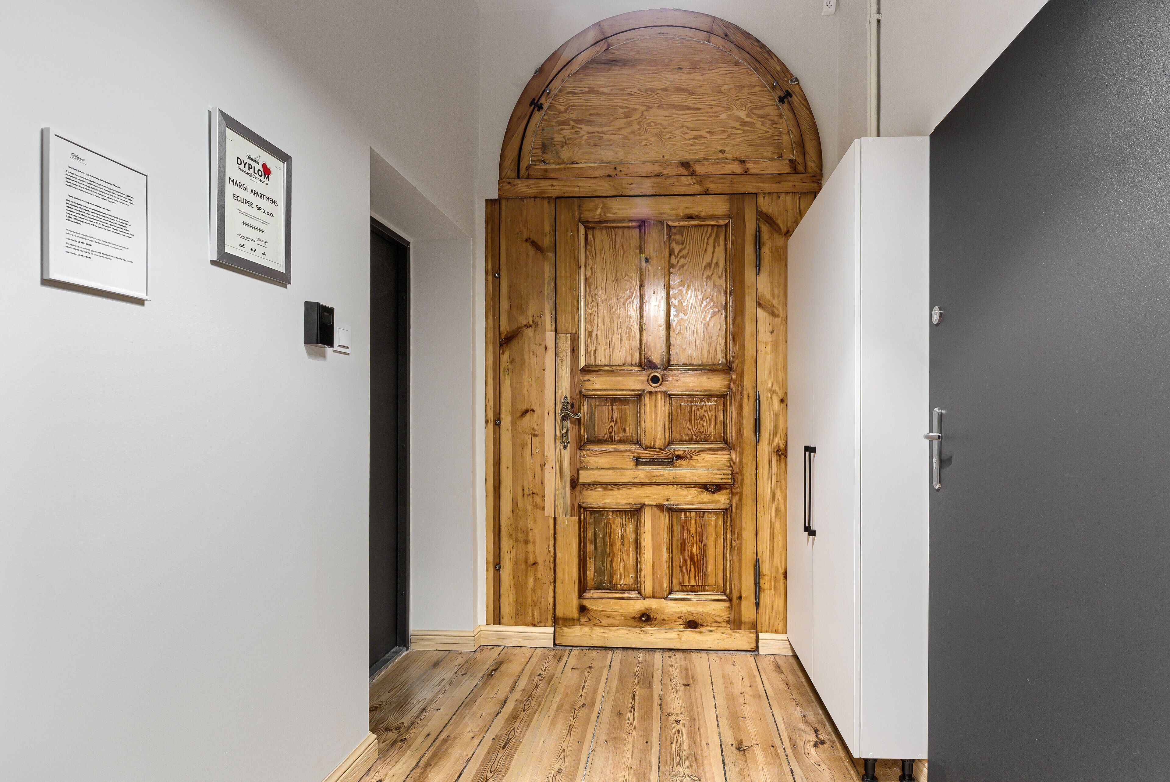 Una puerta de madera con diseño rústico, una pared blanca con diplomas enmarcados y una puerta gris a la derecha.