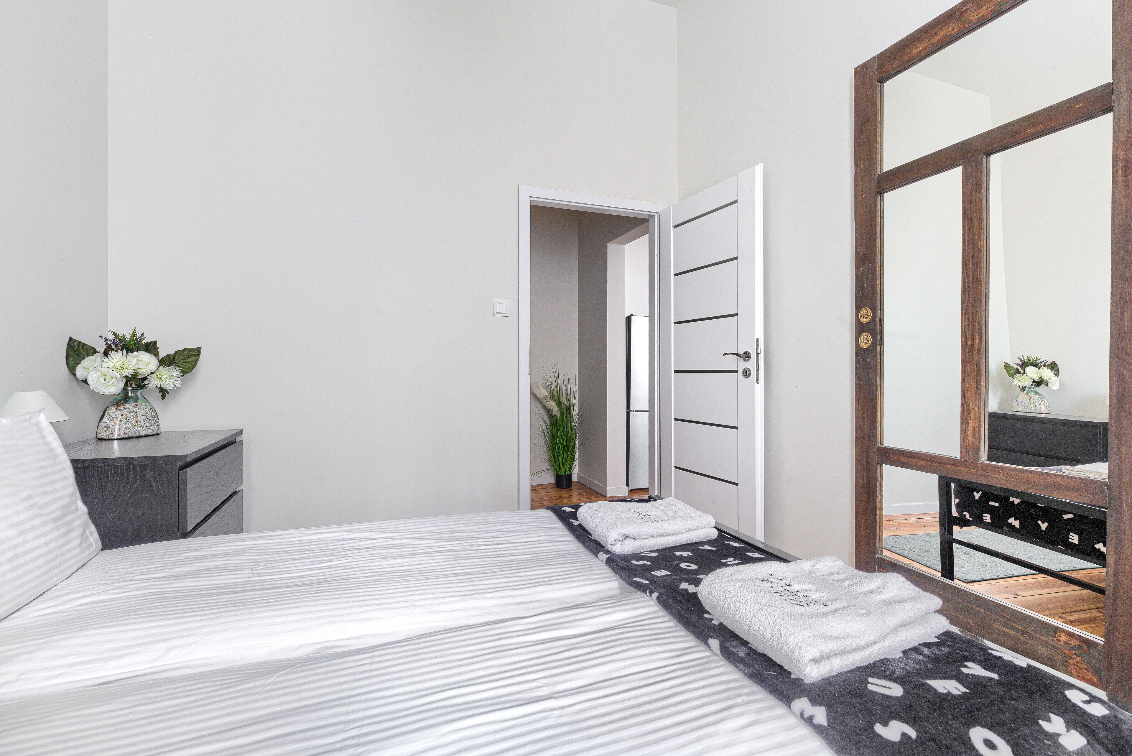 A modern bedroom with a bed, a nightstand, a mirror, and a door leading to another room.
