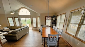 A spacious living area with a wooden dining table, white chairs, a leather sofa, and large windows with blinds. - CP760- Crown Point Home with a Hot Tub and Oceanside! (Corolla)