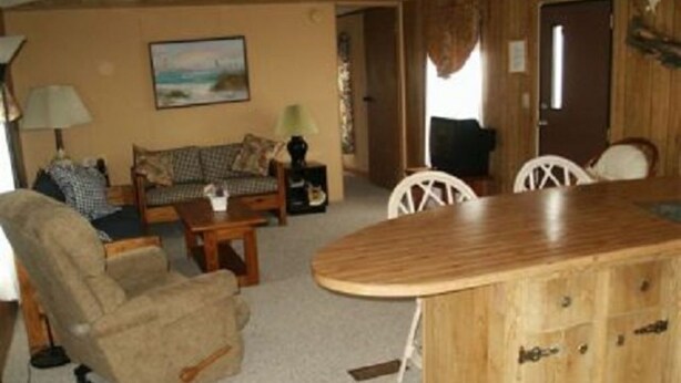 A cozy living room with a sofa, armchair, and wooden coffee table. There is a dining table with chairs and a kitchen area with cabinets.