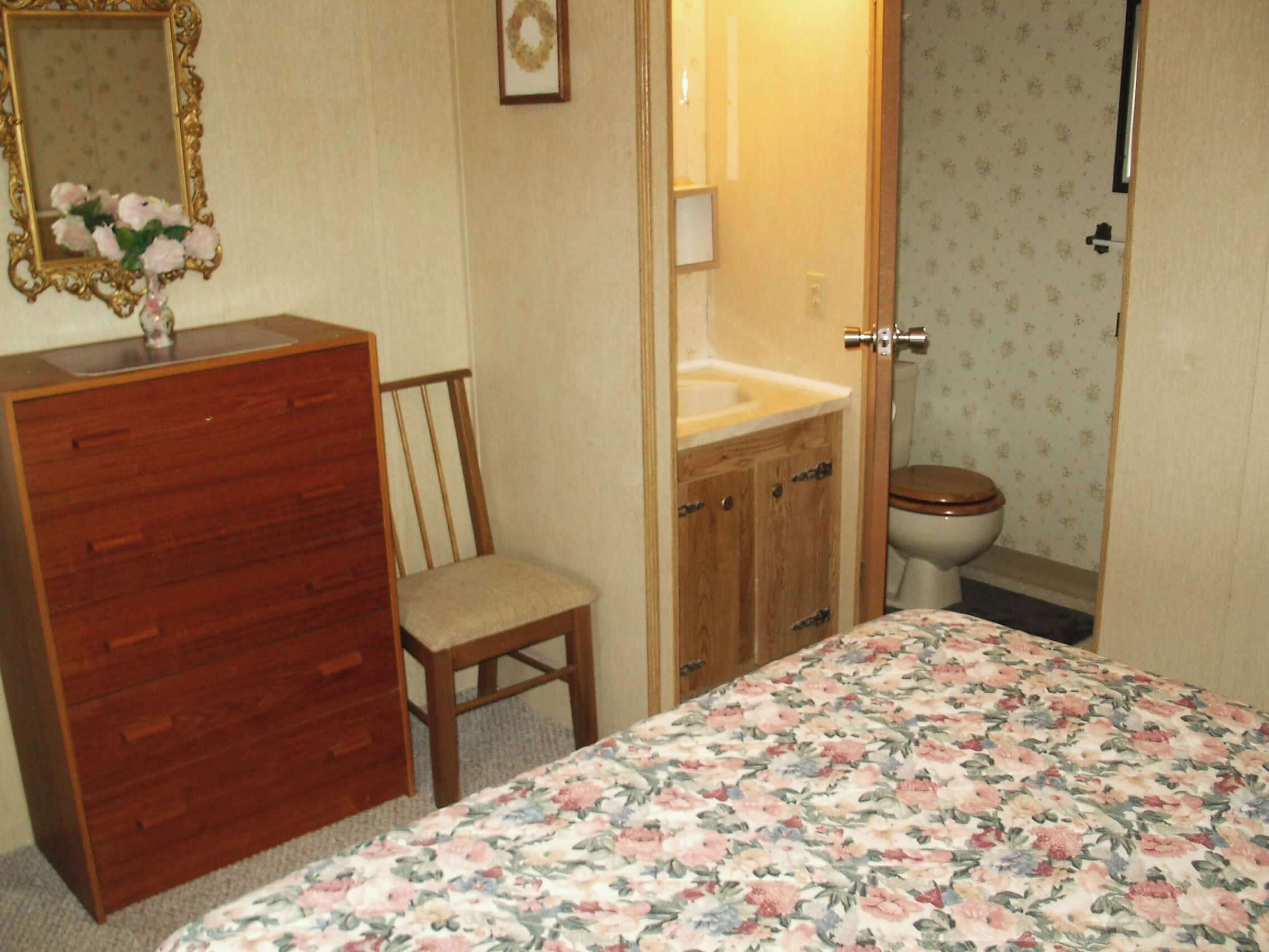 Een slaapkamer met een houten dressoir, een stoel, een bed met bloemenbeddels, een kaptafel met spiegel en een toilet.