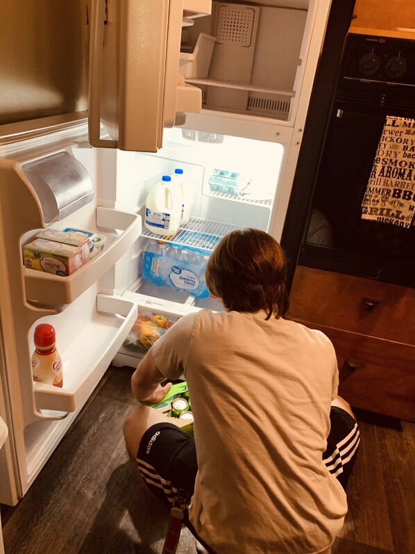 A person sitting on a stool in front of an open refrigerator with milk, butter, and bottled water.