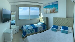 Een slaapkamer met een groot bed, een kleiner bed, een televisie, een dressoir en een raam met uitzicht op zee.