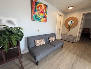 A living room with a grey sofa, a colorful abstract painting, a woven wall hanging, and a large green plant. - Historic Cozy Apartment near Hospital & VSU -SF (Valdosta)