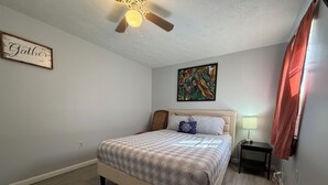 Une chambre à coucher avec un lit, un ventilateur de plafond, un tableau et une table de chevet avec une lampe.