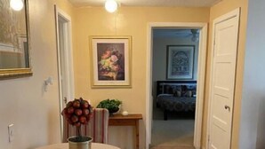Un couloir avec une table ronde et des chaises, une porte sur la gauche, et une chambre visible à travers une porte ouverte au fond.