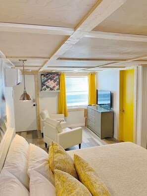 Une chambre d’hôtel avec un lit, un fauteuil blanc, un bureau avec une télévision et une fenêtre avec des rideaux jaunes.