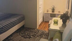 Um quarto pequeno com cama, mesa de cabeceira com uma planta, cadeira e escrivaninha com abajur.