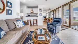 A spacious living area with a sofa, a coffee table, and a leather chair. There is a kitchen in the background with a dining area.
