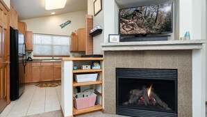 Een woonkamer met een open haard, een televisie erboven en een keukenhoek met houten kastjes en een raam met zonwering.