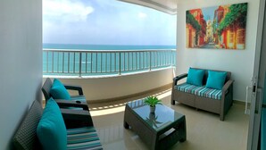 A balcony with a view of the ocean, a sofa with striped cushions, a small table with a plant, and a painting on the wall.
