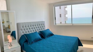 A bedroom with a large bed, a bedside table with a lamp, a mirror, and a view of the sea through a window.