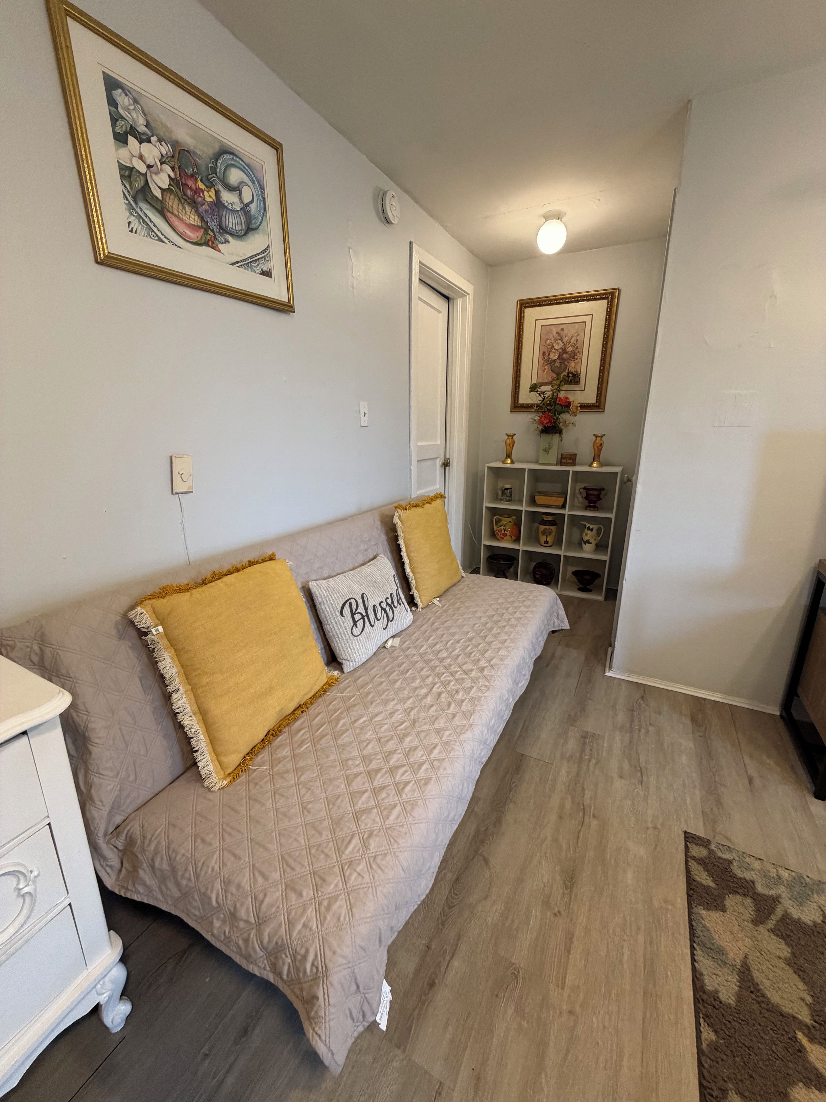 Une pièce avec un canapé beige, une commode blanche, une petite étagère ornée d’objets décoratifs et un tableau encadré accroché au mur.