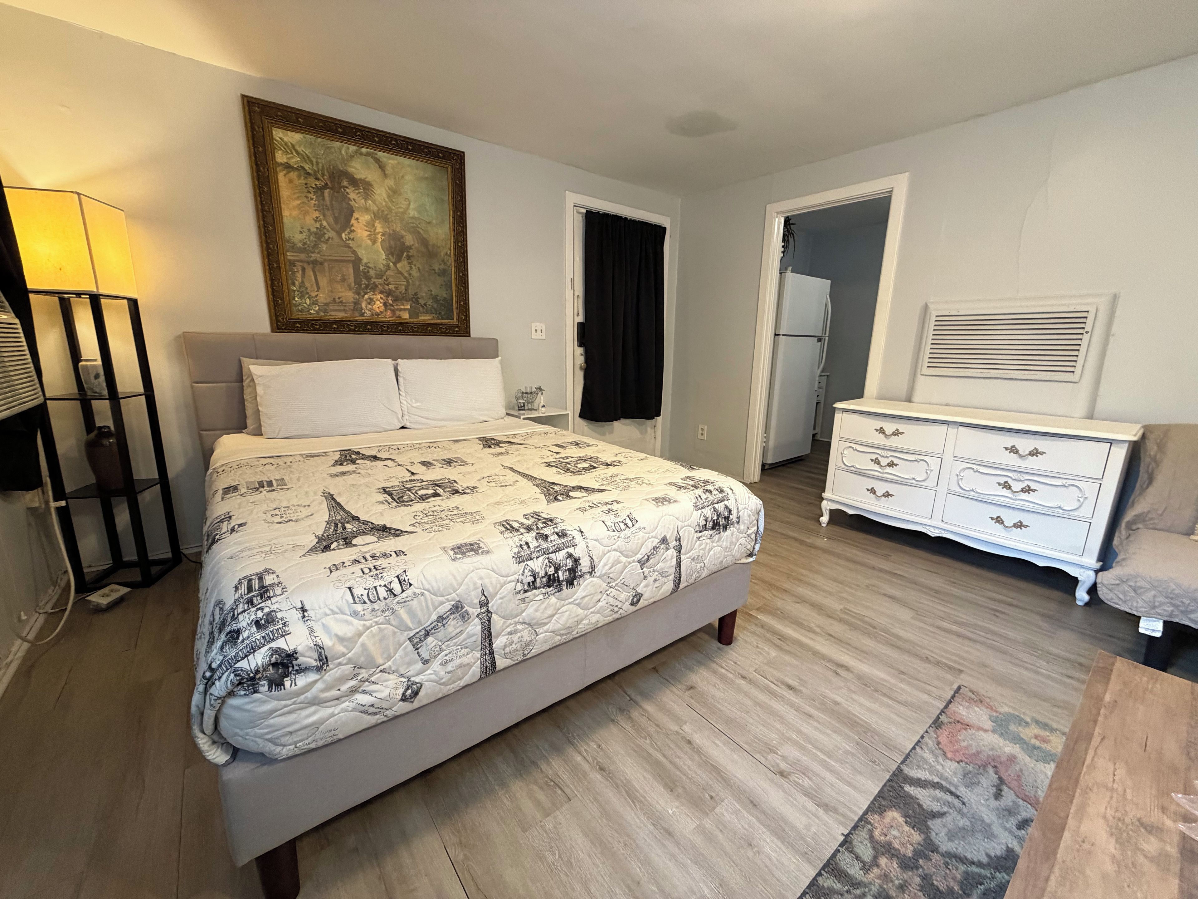 A bedroom with a bed, bedside table, lamp, dresser, and a painting on the wall.